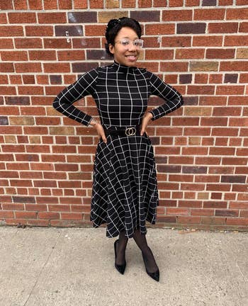 Reviewer wearing high-neck black and white long sleeve plaid dress with black and gold belt, black tights, and black pointed toe heels, standing in front of brick wall