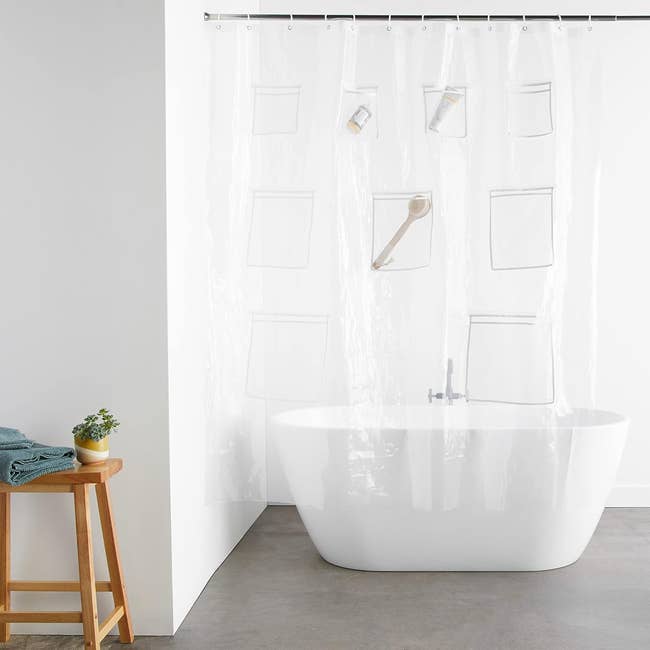 Clear shower curtain with pockets for storage hanging over a white bathtub; nearby stool holds folded towels and a plant