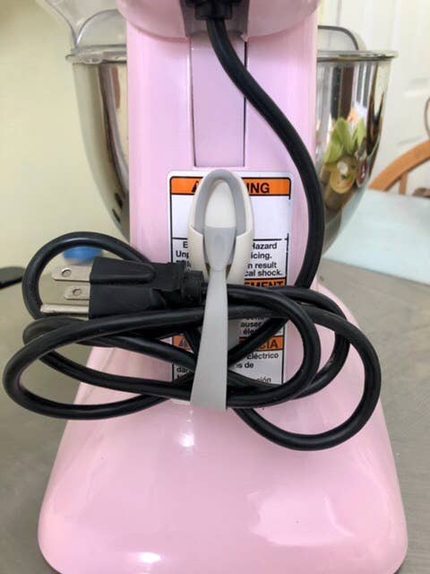 A stand mixer with a pink base has its black power cord neatly wrapped and secured with a white strap, ready for kitchen use