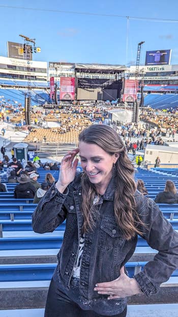 Person smiling and posing in a stadium with a stage set up for a concert in the background