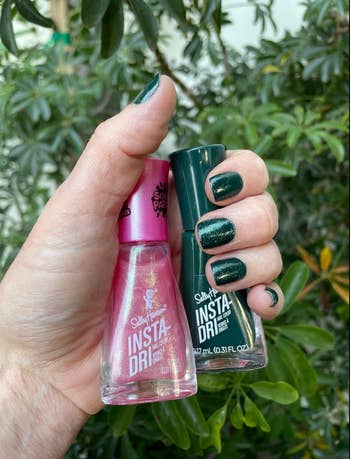 A hand with green-painted nails holds two nail polish bottles, one pink and one green, against a leafy background