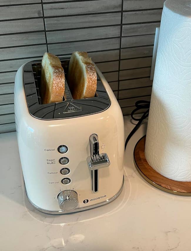 A two-slice toaster with bread and settings for bagel, muffin, reheat, and defrost, next to a paper towel holder on a kitchen counter