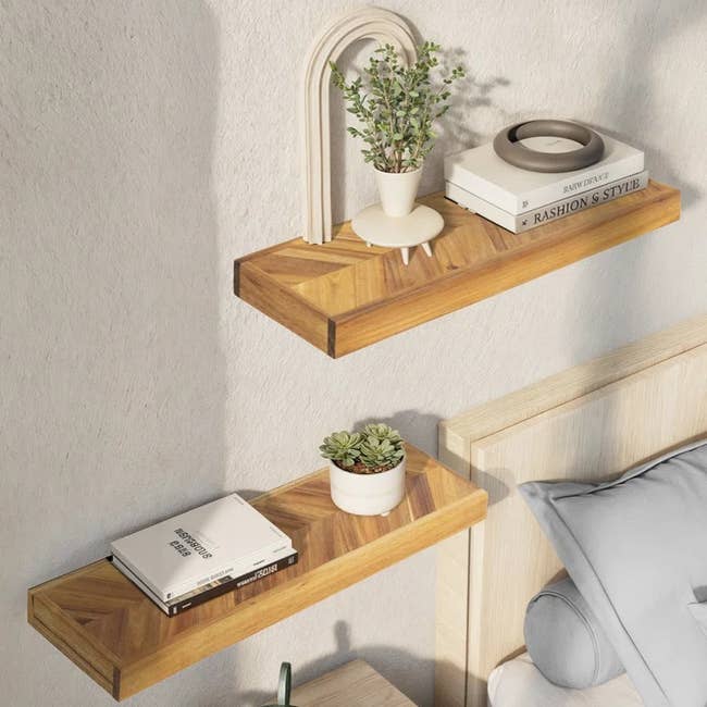 Two wooden wall shelves: top holds books, a potted plant, and decor; the bottom has a small plant and books. Bed corner is visible