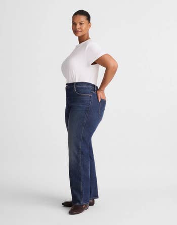 A model posing in a fitted white t-shirt, dark blue jeans, and brown shoes, shot from the side with hands on hips
