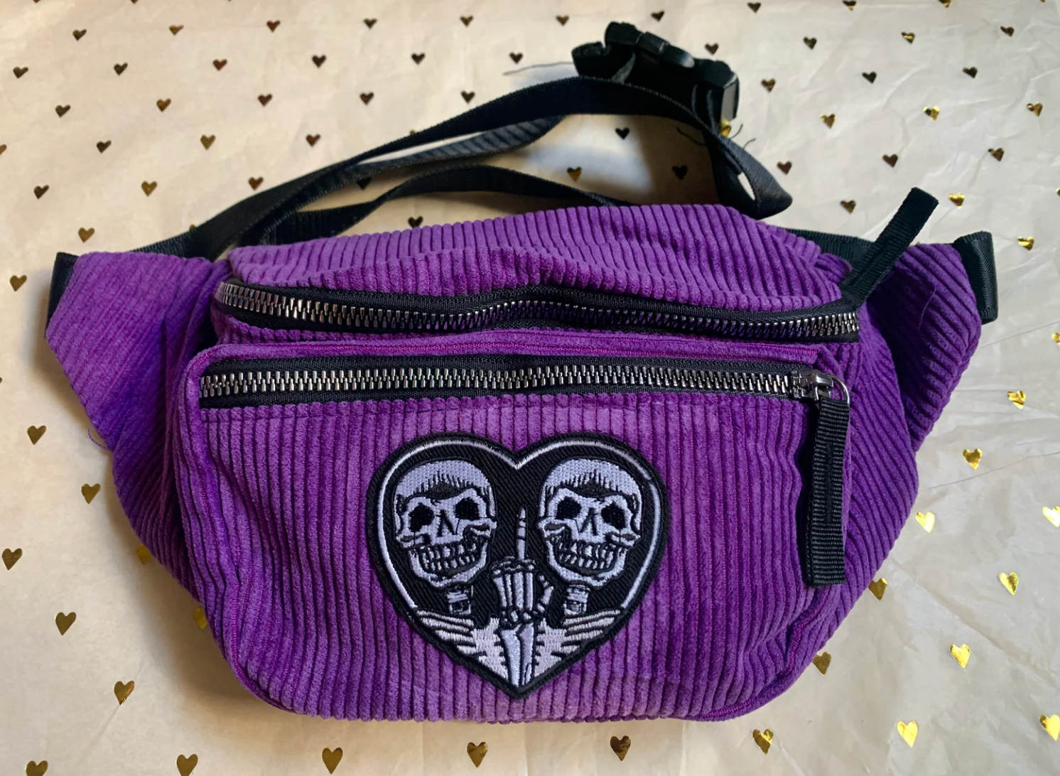 Purple corduroy fanny pack with two zippers and heart-shaped patch with two skeletons on it 