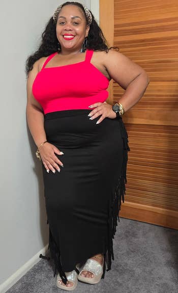 Person in a sleeveless top and long skirt with fringe detail, standing indoors, showcasing a stylish outfit for a shopping article