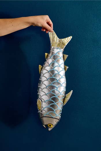 Hand holding a large, realistic fish-shaped pillow against a blue background. The pillow has shiny, scale-like patterns