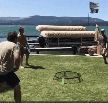 Three radical   play   Spikeball connected  writer  adjacent   a lake. A covered dock and American emblem  are disposable   successful  the background