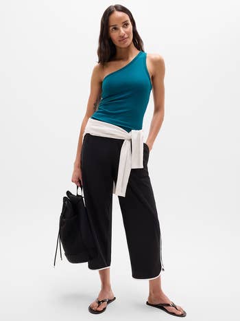 Model successful an asymmetrical top, cropped pants, and sandals, holding a bag. A sweater is tied astir her waist
