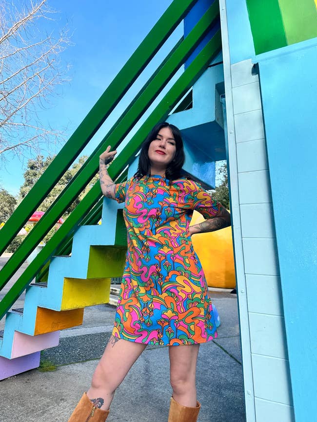 Woman posing in a vibrant patterned dress and brown boots near a colorful staircase