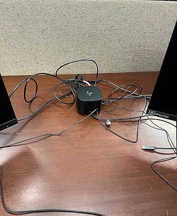 Messy desk with tangled cables connected to a small electronic device between two monitors and a keyboard