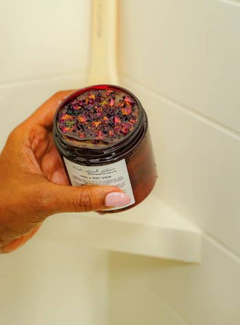 Hand holding a jar with a purple exfoliating scrub by a bathroom sink, suggesting skincare shopping