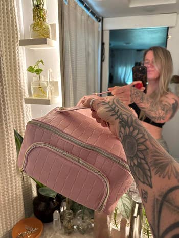 Orang bertato memegang tas kosmetik bertekstur dalam selfie cermin di lingkungan rumah