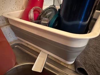 Collapsible dish rack draining by a kitchen sink, holding assorted tumblers and lids, ideal for compact storage and efficient drying solutions