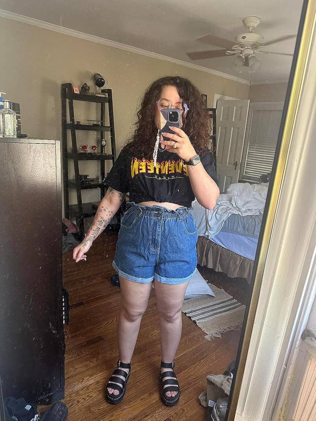 Person taking reflector selfie wearing casual outfit with a graphic tee and denim shorts, showcasing a stylish, summery look suitable for shopping