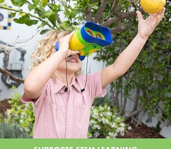 A child looking through the binoculars while reaching for a fruit hanging from a tree