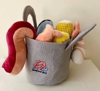 Fabric bucket with plush seafood toys, including shrimp, corn, and crab claw, labeled 