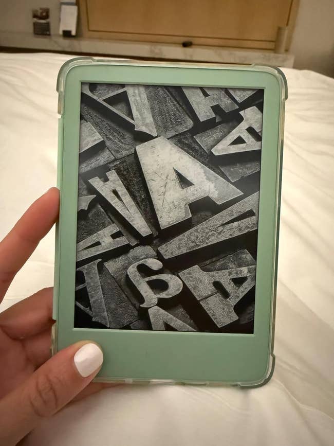 A hand holding an e-reader displaying a black-and-white image of scattered letterpress typefaces, featuring a prominently large letter 