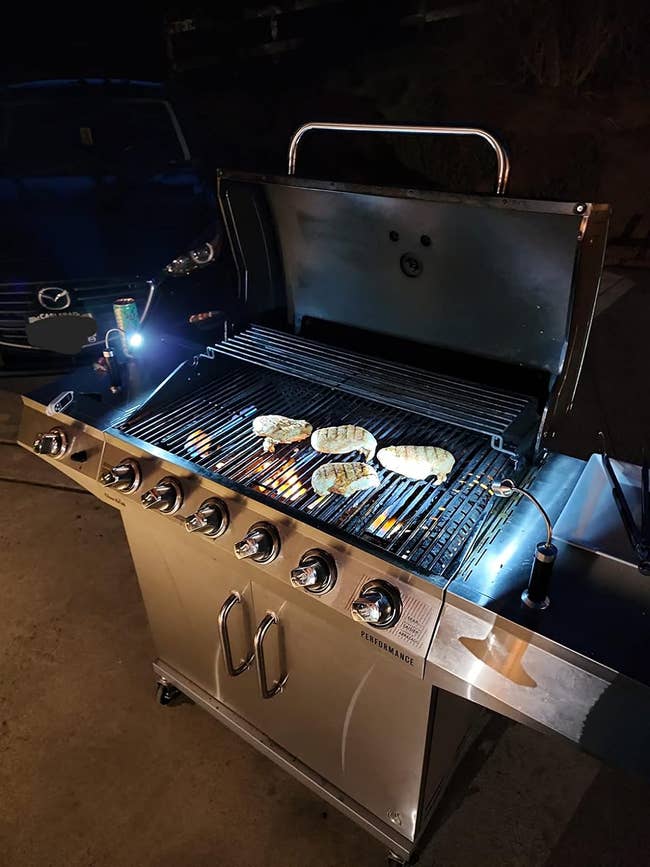 A gas grill with six burners cooking four pieces of meat at nighttime, with a car in the background