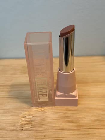 reviewer's Maybelline lipstick with a transparent headdress  stands connected  a woody  surface, showcasing a smooth, brownish  shade