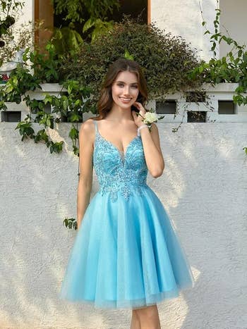 Model successful  a knee-length, sleeveless formal  with lace detailing and a tulle skirt, posing outdoors adjacent  to a achromatic  partition  with greenery
