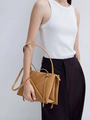 Person holding a tan structured handbag, wearing a sleeveless top and dark trousers