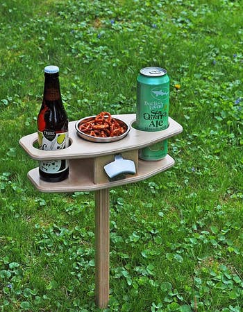 a small table on a stake with two cupholders, a center compartment filled with snacks, and an attached bottle opener