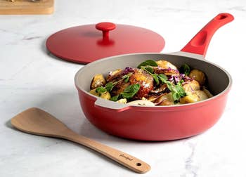 Red non-stick frying pan with lid, containing cooked vegetables, accompanied by a wooden spatula