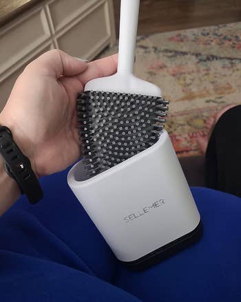 Reviewer holding a flat silicone head gray toilet brush that fits into a white holder 
