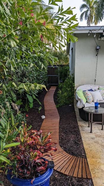 A backyard scene showing a narrow wooden walkway surrounded by lush greenery, leading to a fenced gate. A patio area to the right features a white cushioned seat and a small table with a blue drink tumbler