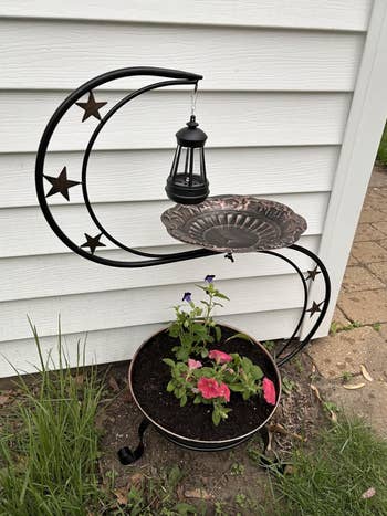 Decorative plot  planter with a crescent-shaped stand, prima  accents, and a hanging mini lantern, holding flowers and a crockery  above, adjacent  to a wall