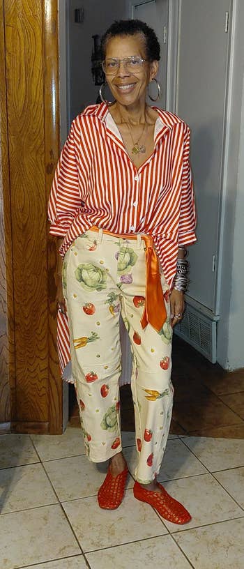 Person successful a reddish and achromatic striped garment with floral patterned pants and reddish shoes, posing indoors for a buying article