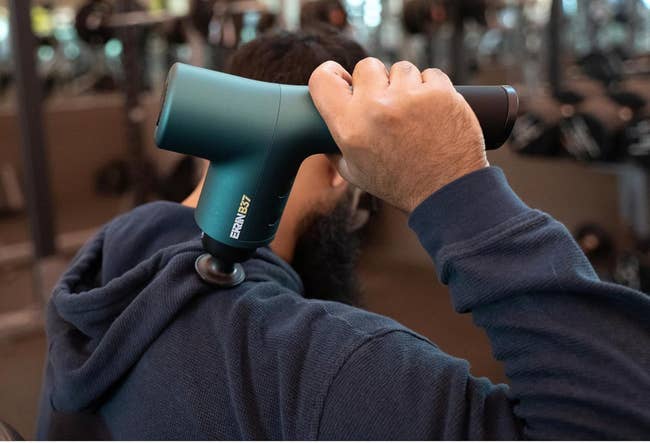 Person using a handheld massage gun on their shoulder in a gym setting, focusing on personal wellness and recovery