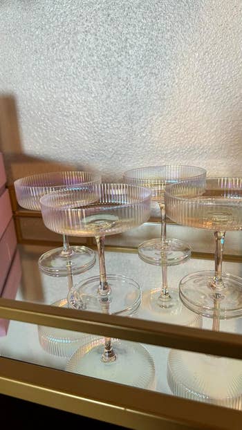 Four elegant glass champagne coupes displayed on a reflective surface beside pink gift boxes