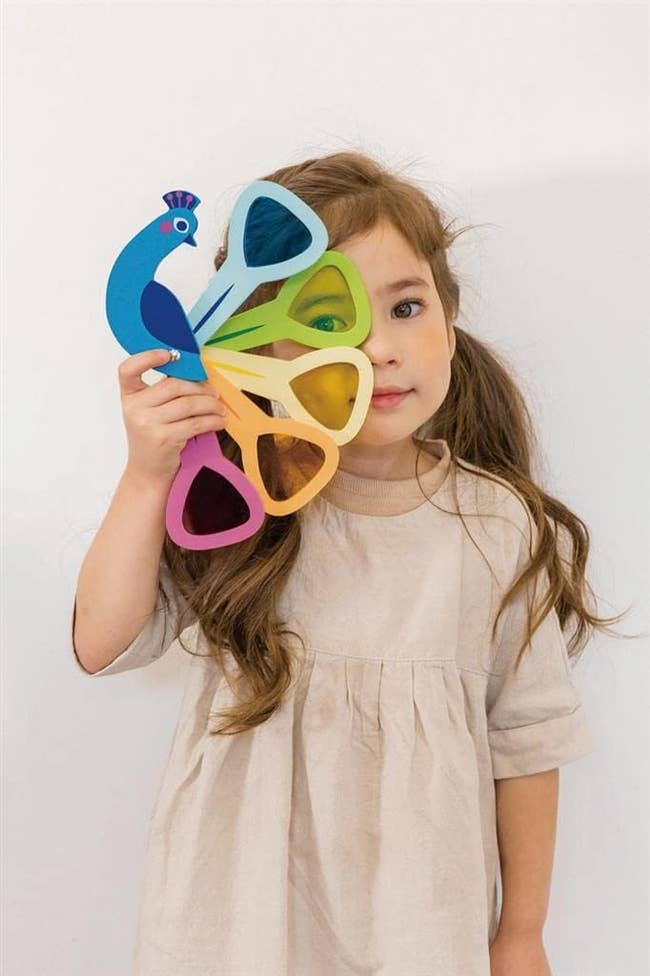 Child holding a fan-shaped toy with peacock design and colorful cutouts, standing against a plain background