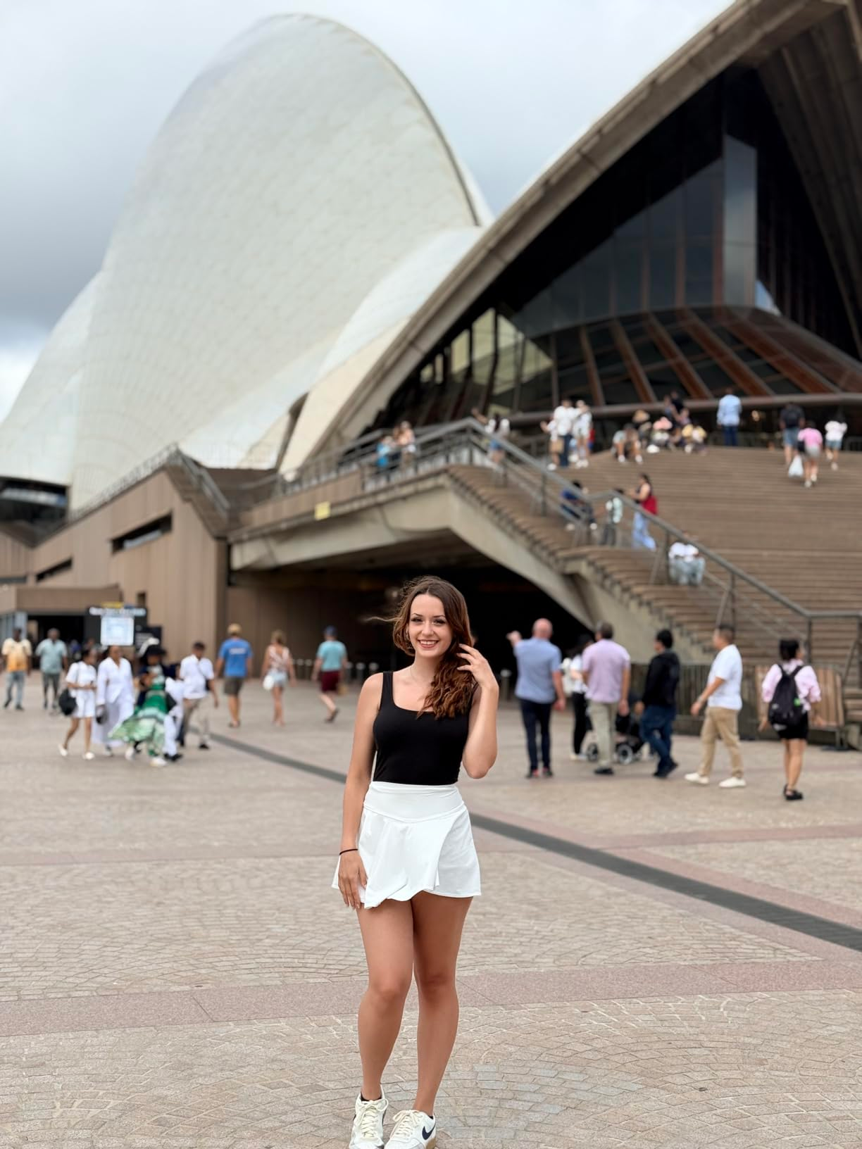 Person successful  a casual achromatic  apical  and achromatic  skirt stands smiling successful  beforehand   of the Sydney Opera House, with radical   walking successful  the background
