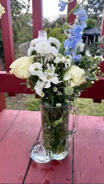 A bouquet of mixed flowers including white roses, blue blossoms, and various greenery arranged in a glass boot-shaped vase, set on a wooden surface