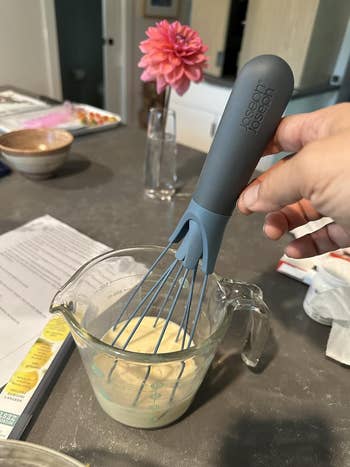 Hand holding a whisk wrong  a measuring cupful  with batter, connected  a room  counter. A pinkish  angiosperm  successful  a vase is successful  the background