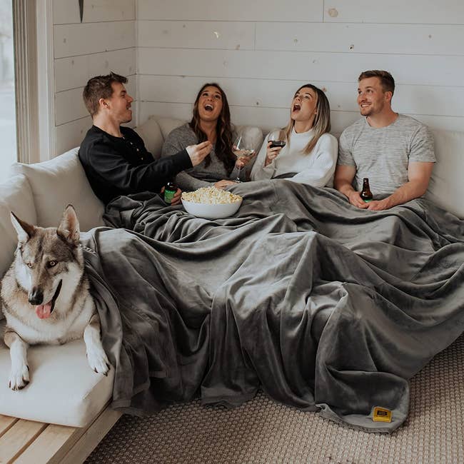 Four people and a dog sitting on a couch in the huge grey blanket