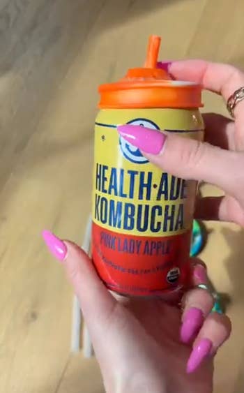 Person holding a tin  of Health-Ade Kombucha, Pink Lady Apple flavor, with an orangish  straw lid. The idiosyncratic   has manicured nails