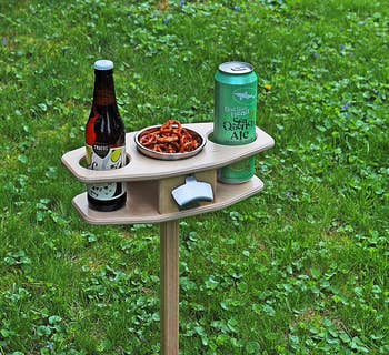 a small table on a stake with two cupholders, a center compartment filled with snacks, and an attached bottle opener 