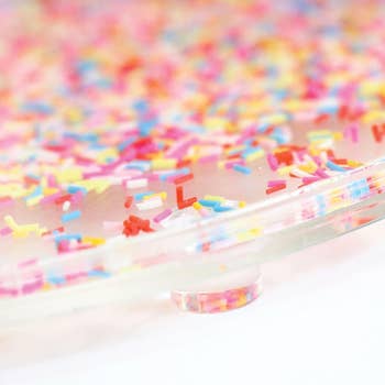 Close-up of a clear cake stand sprinkled with multicolored confetti, likely used to display festive desserts