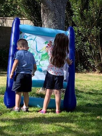 Two children painting on an upright inflatable canvas outdoors in a grassy area
