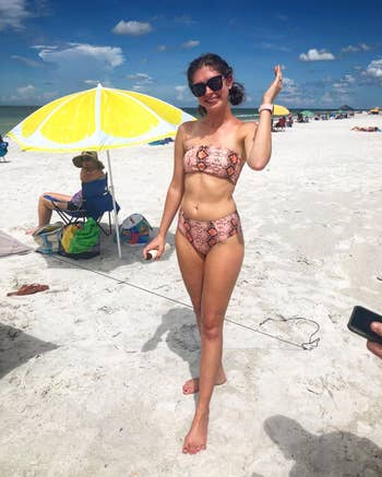 Person on a beach wearing a patterned bikini, waving. Umbrella and other beachgoers in the background