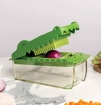 Vegetable chopper with a crocodile-themed design, featuring a green lever and blade for slicing onions and other produce in a clear container