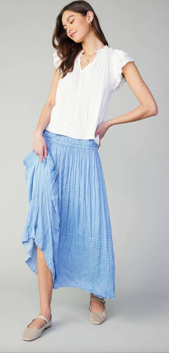 Woman in white blouse and blue pleated skirt with hand on hip, looking down. Item likely for sale
