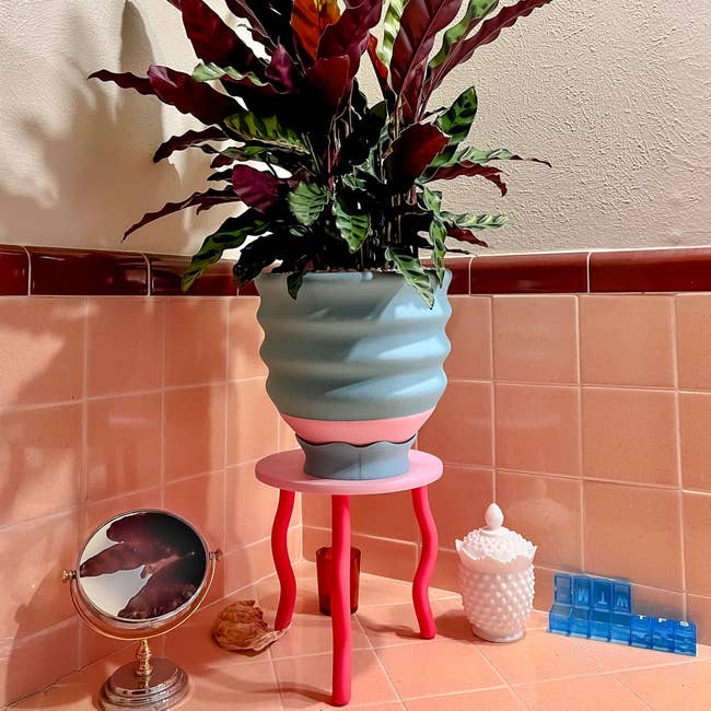Decorative works successful a wavy vase sits connected a pinkish stool successful a tiled corner, accompanied by a tiny circular reflector and a achromatic decorative container