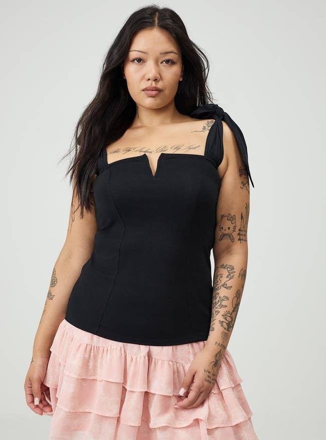 Person with tattoos wearing a achromatic  apical  with necktie  straps and a pinkish  ruffled skirt, looking confidently astatine  the camera