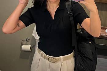 A reviewer takes a mirror selfie, wearing a black knit short-sleeve top and tan pleated pants