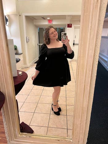 reviewer posing in a mirror wearing a black, long-sleeved, knee-length dress with a square neckline and black heels, taking a selfie